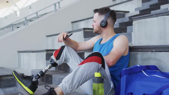 Caucasian disabled male athlete with prosthetic leg sitting, wearing headphones, using smartphone