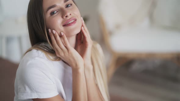 Beautiful Young Woman Sitting in Bedroom on Bed, Looking at Camera, Smiling