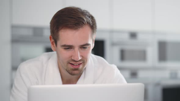 Close Up Portrait of Young Smiling Man in Bathrobe Reading Email on Laptop in Kitchen