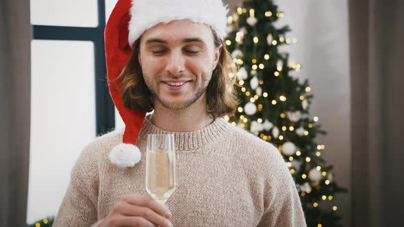 Young Man in Santa Hat is Drinking Champagne and Smiling While Standing at Home Against Decorated