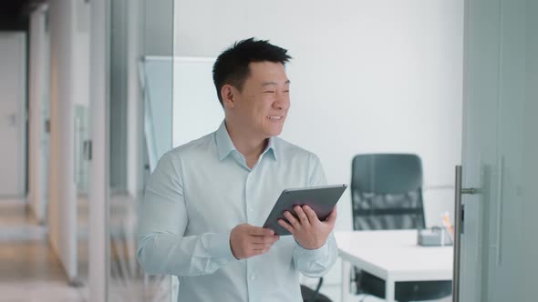 Positive Middle Aged Asian Businessman Reading Social Media on Digital Tablet and Smiling at Office