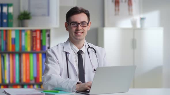 Portrait of Happy Male Doctor in White Coat and Stethoscope Smiling at Camera Sitting at Desk