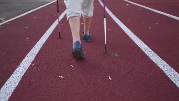 Closeup of a Senior Woman Legs Walking on a Stadium Track with Nordic Walking Poles Rear View