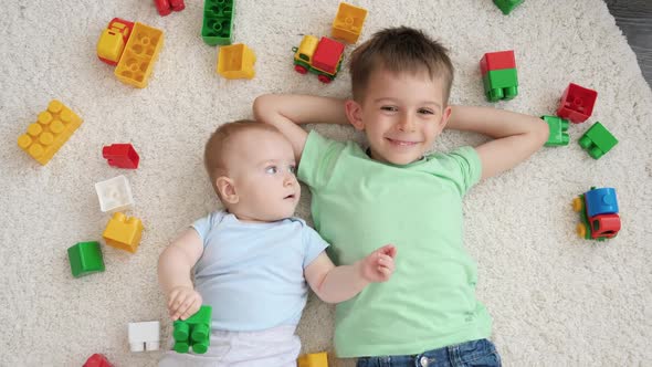 Smiling Baby Boy with Older Brother Lying on Carpet Next to Heap of Colroful Toys and Looking Up in