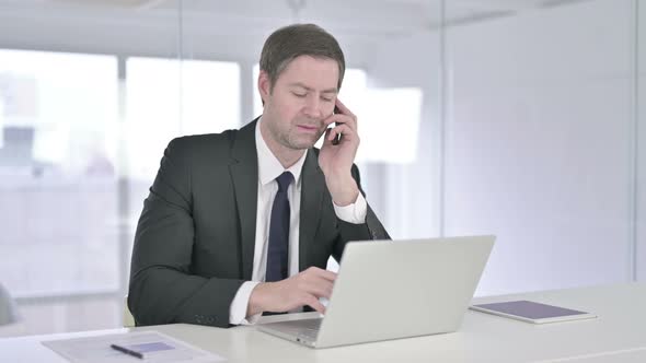 Middle Aged Businessman Talking on Smartphone and Working on Laptop