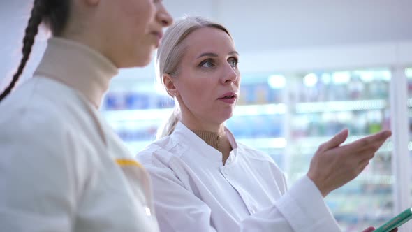 Beautiful Expert Female Druggist with Tablet Standing at Shelves in Pharmacy Talking with Blurred