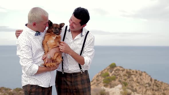 Two Stylish Cheerful Man with Cute Dog Sitting on Stones Along Beach and Talking Happy Gay Couple