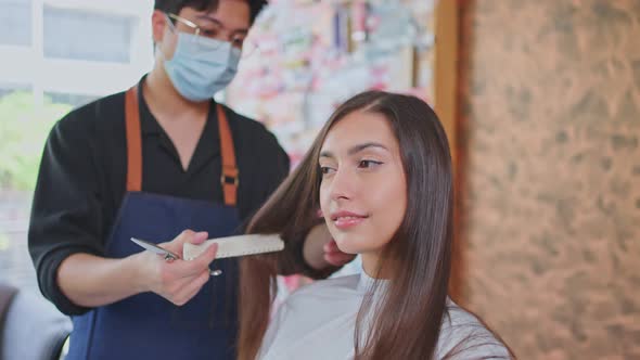 Asian male hairdresser hairstylist wear mask, using scissors cutting young customer's hair in salon