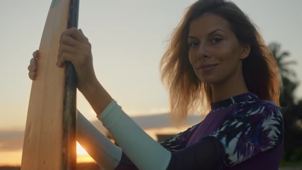 Young Attractive Female Surfer Smiling and Posing with Her Surfboard at Sunset on the Beach