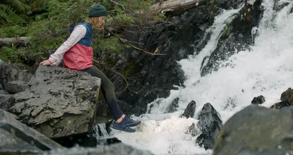 Young Woman Sitting on Rock Above Creek Waterfall in Wilderness of Alaska, USA. Cinematic Slow Motio