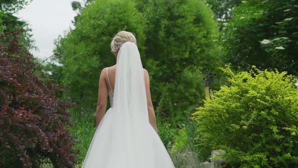 A Young Blonde in an Elegant Long White Wedding Dress with a Long Train Stands with Her Back to the