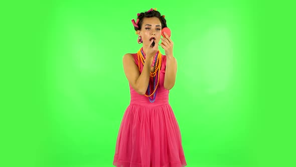 Girl with Curlers on Her Head in a Pink Dress Paints Her Lips Looking in Red Mirror. Green Screen