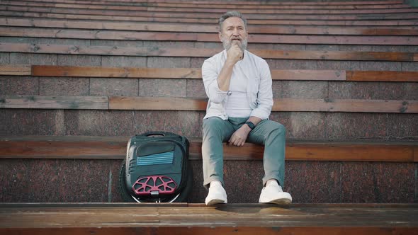 Elderly Man Scratches Beard Sitting on Stairs in City Park