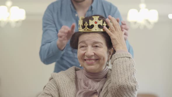 Portrait of Cute Pretty Mature Woman. Adult Grandson Brings the Crown and Puts It on the Head of