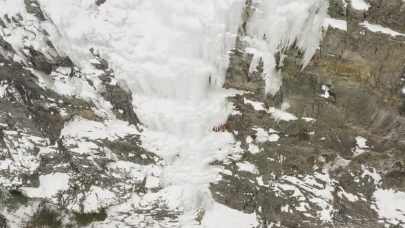 Sheer cliff frozen cascade climbers under overhang on ledge Aerial 4K