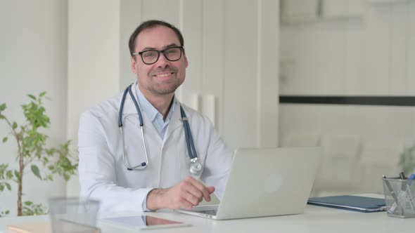 Middle Aged Doctor with Laptop Smiling at the Camera in Office