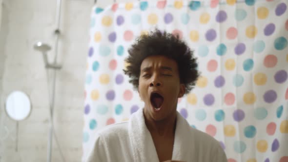 Portrait of Young African Guy in Bathrobe Yawning and Stretching at Bathroom in Morning