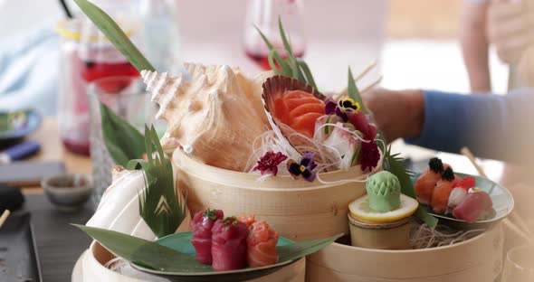 Person Picking Fresh Sushi On A Platter Decorated In Seashells, Flowers, And Green Leaves. - close u