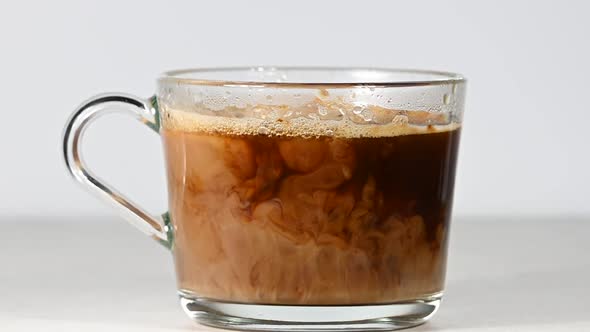 Pouring milk in glass cup of black coffee over white background