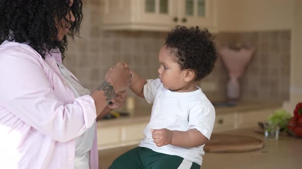 Curios African American Boy Sitting on Countertop at Home Watching Mother Preparing Children Food in