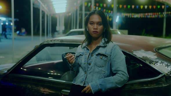 a Sad Thai Girl Sits Smoking Leaning Against a Rusty Car at Night