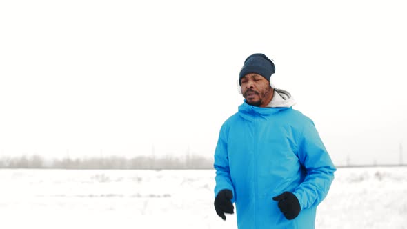 Black man at jacket, hat and headphones is jogging in winter outdoors, front view. African-American