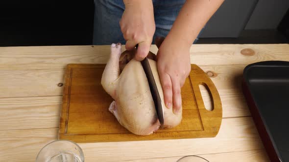 A woman is cutting a raw chicken carcass in half on a cutting board with a large knife