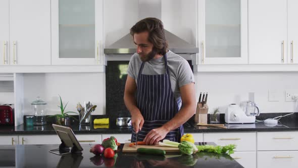 Caucasian man wearing apron chopping vegetables while looking at digital tablet in the kitchen