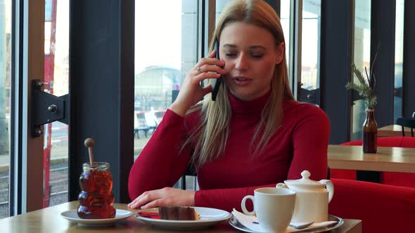 A Young Attractive Woman Sits at a Table with Meal in a Cafe and Talks on a Smartphone with a Smile