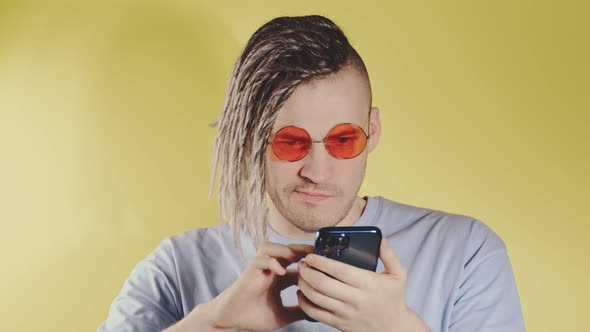 Young Disappointed Puzzled Man in Bright Sunglasses Browsing Mobile Phone on Yellow Background