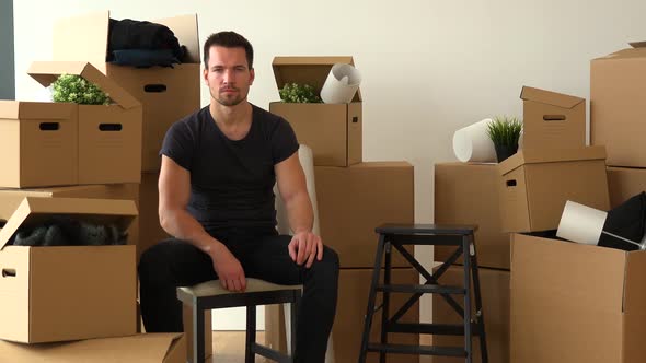 A Moving Man Sits on a Chair and Looks Seriously at the Camera in an Empty Apartment