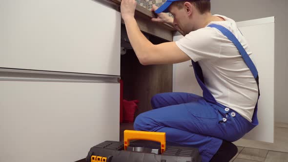 plumber in overalls and white T-shirt examines pipes in kitchen in apartment.