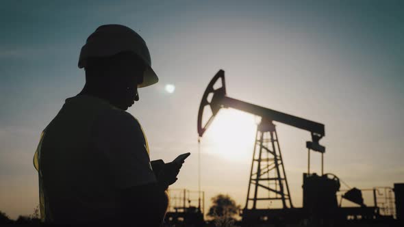 Silhouette of Man Engineer with Phone Overseeing the Site of Crude Oil Production at Sunset.