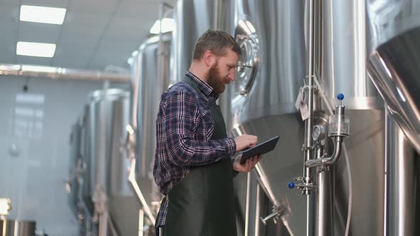 Brutal Male Brewer with a Beard Is Standing Near the Beer Tanks and Captures the Readings Using a