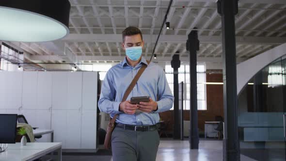 Caucasian businessman walking through corridor wearing facemask holding tablet