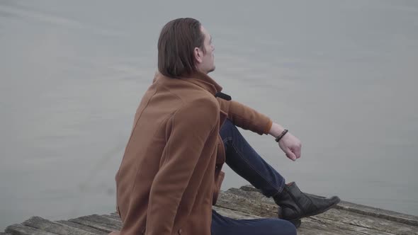 Confident Young Man Sitting on Pier and Thinking. Side View of Elegant Brunette Caucasian Guy in