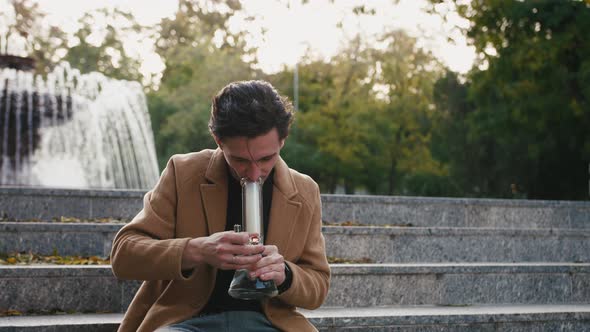 Handsome Stylish Young Man Sitting on Stairs and Smoking Weed with Bong in Park