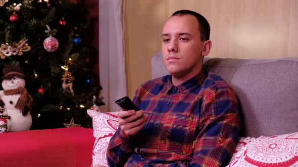 Portrait of a Lonely Man Sitting on Sofa Near the Christmas Tree and Watching Tv