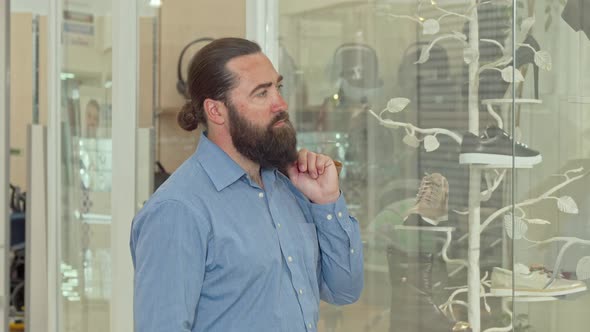 Mature Bearded Man Looking Thoughtfully at the Display of Clothing Store