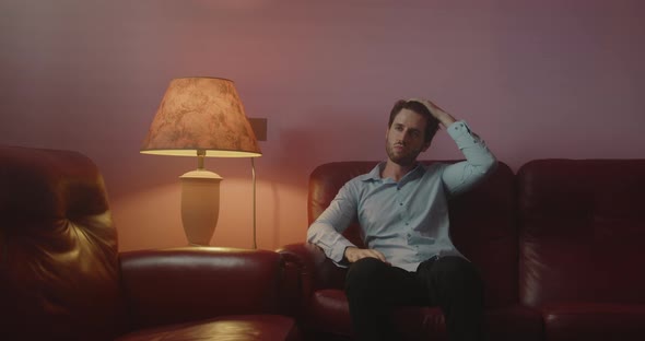 Nervous guy touches his beard while sitting on the sofa at home