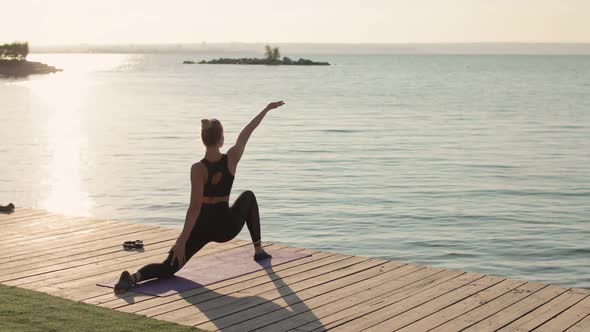 Woman Is Practicing Yoga Doing Lunge Warrior Yoga Pose on River Pier, Back View.