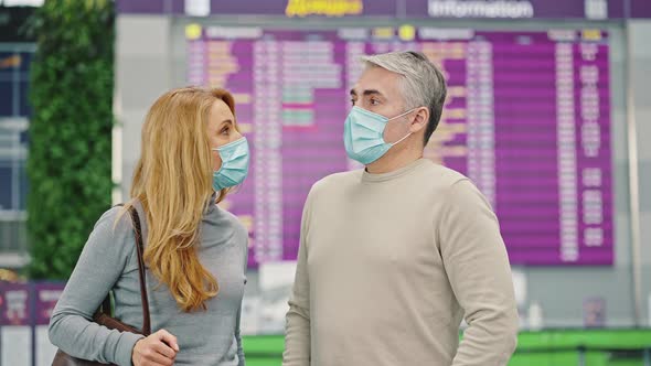 Middle Aged Spouses Wearing Protective Medical Masks Standing Near Time Board at Airport Getting