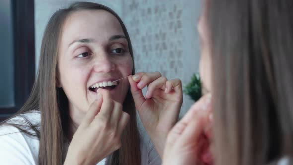 Young Woman Brushes Teeth with Dental Floss in the Bathroom Mirror Reflection