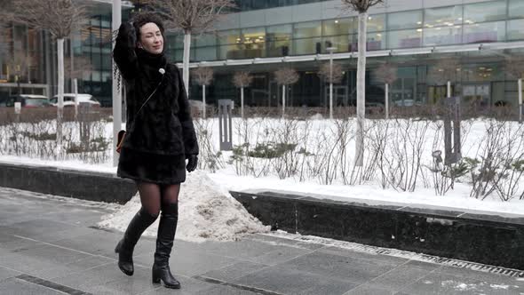 A Curlyhaired Woman in a Fur Coat Walks Through a Modern City Enjoying a Beautiful Day