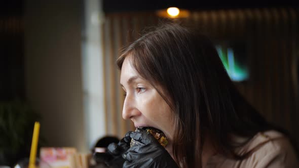 Side View of Young Girl in Black Gloves Eating a Burger in Cafe