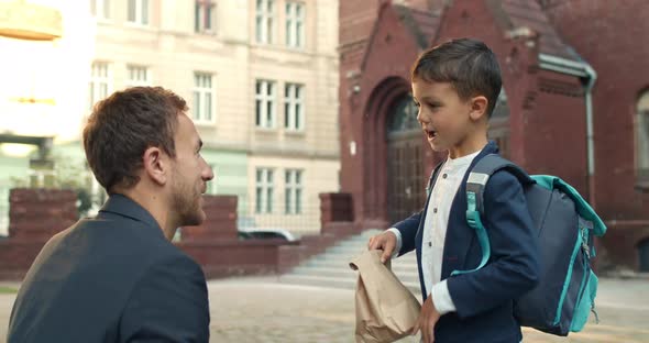 Young Man Giving Lunch To His Kid Boy While Sending Him To School. Male Child with Backpack Giving