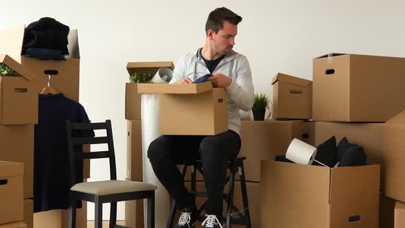 A Moving Man Sits on a Chair and Takes Things Out of a Cardboard Box in an Empty Apartment