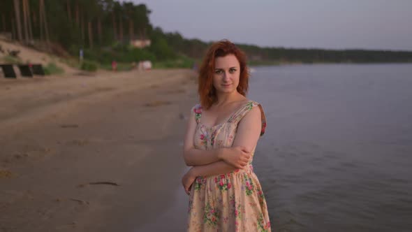 Dolly in Pretty Young Woman in Summer Dress Stands at River Coast Looking at you at Sunset