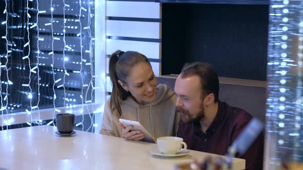 A couple sitting in a cafe, talking, drinks coffee and watching in smartphone.