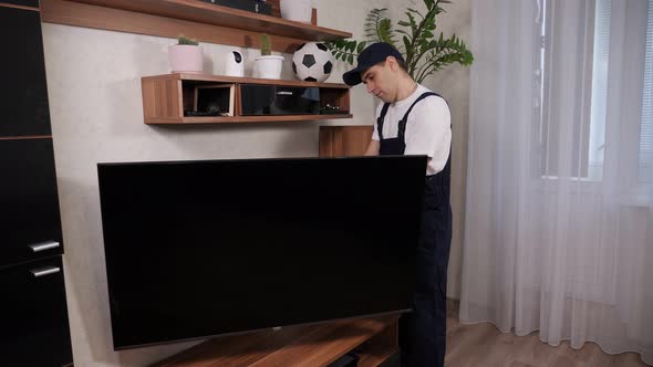 A Male Technician in Blue Overalls Repairs a TV at a Customer's Home